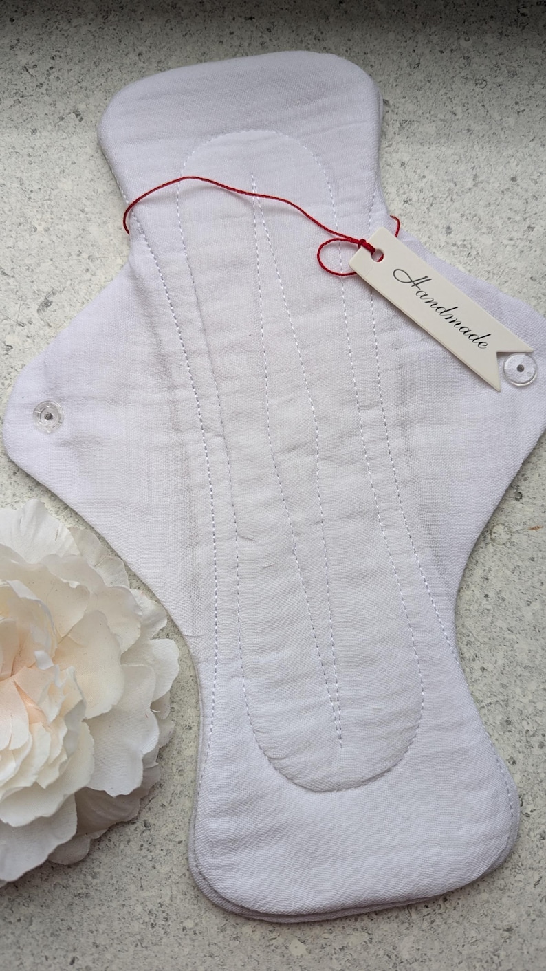 May include: A white cloth menstrual pad featuring vertical quilted stitching lines and two clear plastic snap buttons on the side wings. A small off-white tag with the word Handmade printed in black is attached by a thin red string.