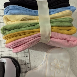 May include: A stack of six folded fabric pads in black, light blue, yellow, green, mustard, and pink, secured by a cream-colored ribbon with a snap button. A white fabric pad with a yellow stitched flower design sits nearby on a cutting mat.