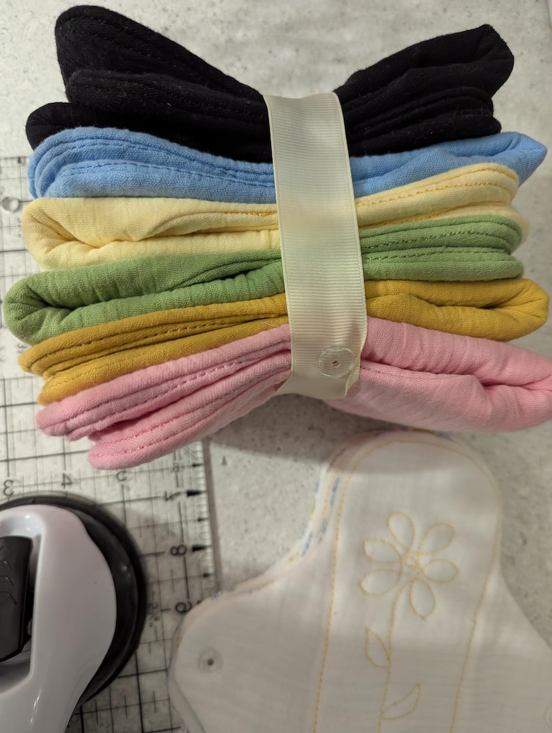 May include: A stack of six folded fabric pads in black, light blue, yellow, green, mustard, and pink, secured by a cream-colored ribbon with a snap button. A white fabric pad with a yellow stitched flower design sits nearby on a cutting mat.