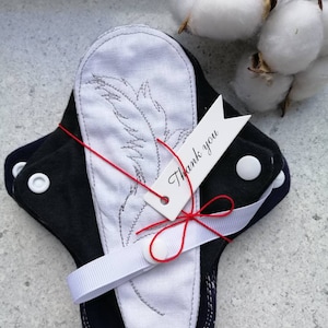 Puede incluir: Un juego de compresas de tela reutilizables de color negro y blanco, atadas con una cinta blanca y una pequeña etiqueta que dice "Gracias".