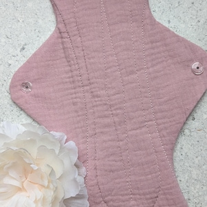 May include: A dusty rose reusable cloth menstrual pad featuring a textured muslin fabric surface with vertical stitched channels and two clear plastic snap fasteners on the side wings, accompanied by a large white artificial peony flower.