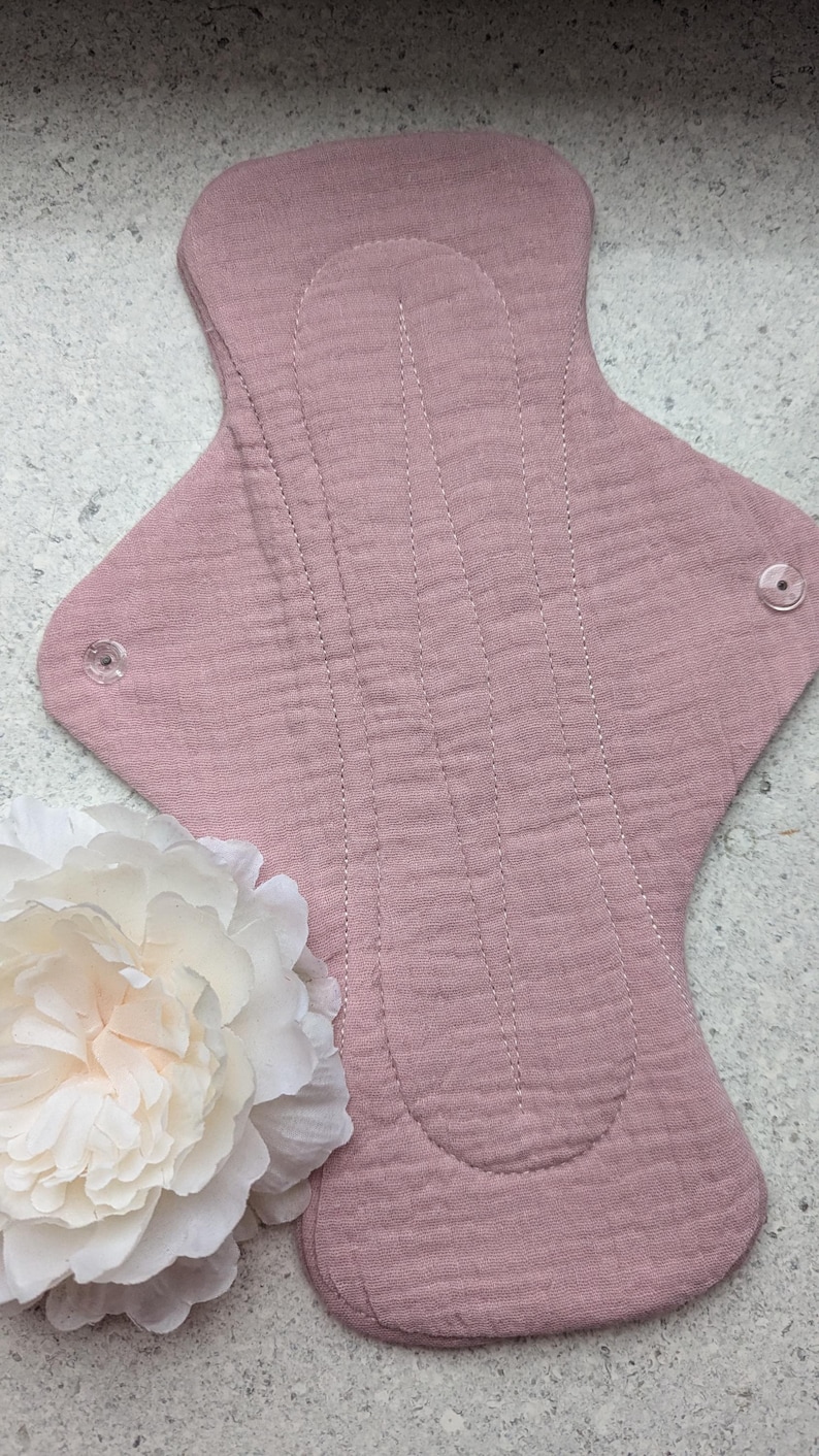May include: A dusty rose reusable cloth menstrual pad featuring a textured muslin fabric surface with vertical stitched channels and two clear plastic snap fasteners on the side wings, accompanied by a large white artificial peony flower.