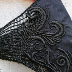 May include: A black fabric face mask featuring intricate black embroidery in a swirling scroll pattern on the side, complemented by a delicate black lace trim along the edge, providing a detailed view of the textile texture and stitching.