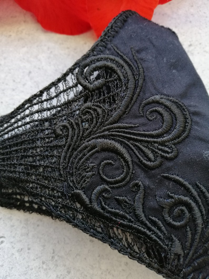 May include: A black fabric face mask featuring intricate black embroidery in a swirling scroll pattern on the side, complemented by a delicate black lace trim along the edge, providing a detailed view of the textile texture and stitching.