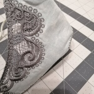 May include: A light blue face mask with a gray lace trim. The lace trim is a delicate pattern of swirls and circles.