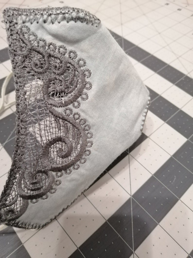 May include: A light blue face mask with a gray lace trim. The lace trim is a delicate pattern of swirls and circles.