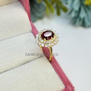 Könnte beinhalten: Ein goldener Ring mit einem großen, ovalen, tiefroten Edelstein, umgeben von zwei Reihen kleinerer, klarer Edelsteine. Der Ring ist in einem goldenen Band gefasst und wird in einer rosa Schmuckschatulle präsentiert. Der Text "ZafiroJewelryCo" ist sichtbar.