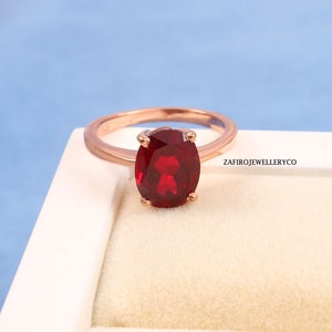 Peut inclure: Une bague en or rose ornée d'une grosse pierre rouge foncé de forme ovale. La bague est posée sur une surface blanche avec un fond bleu. Le texte "ZAFIROJEWELLERYCO" est visible sur le côté droit.