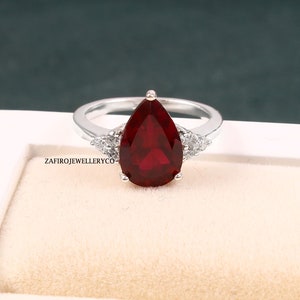 May include: A silver ring with a large, pear-cut, deep red gemstone. The ring is flanked by smaller, clear gemstones. The ring is displayed against a neutral background. The ring is a piece of jewellery.