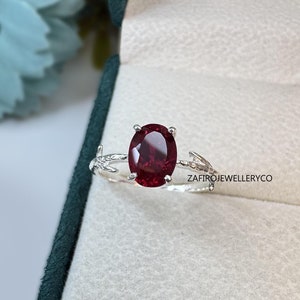 May include: A silver ring featuring an oval-cut deep red gemstone held by four prongs. The band has a textured, branch-like design. The text ZAFIROJEWELLERYCO is visible on the right side of the ring. The ring is set against a light fabric.