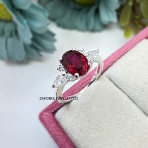 May include: A silver ring featuring a large oval red gemstone, surrounded by smaller clear gemstones. The ring is displayed in a pink velvet ring box.