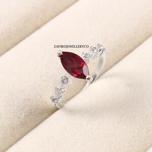 Puede incluir: Un anillo de plata con una gema roja oscura en forma de marquesa. El anillo está adornado con gemas claras más pequeñas a lo largo de la banda. El anillo se presenta sobre un fondo neutro.