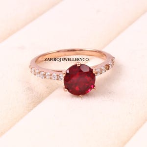 Puede incluir: Anillo de oro rosa con una piedra preciosa grande, redonda y de color rojo intenso. La banda está adornada con pequeñas piedras claras. El texto "ZAFIROJEWELLERYCO" es visible en la banda. Este anillo es una joya.