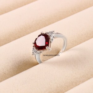 Peut inclure: Une bague en argent avec une grande pierre rouge en forme de cœur et de plus petites pierres claires sur la bande. La bague est sur un fond blanc.
