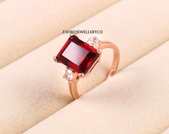 Könnte beinhalten: Roségoldener Ring mit einem großen, rechteckigen, tiefroten Edelstein und zwei kleineren, klaren Edelsteinen. Der Ring hat eine geschwungene Ringschiene. Der Text "ZAFIROJEWELLERYCO" ist sichtbar.