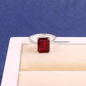 May include: A silver ring with a large, rectangular, red gemstone. The ring is in a white box with a cream-colored interior.