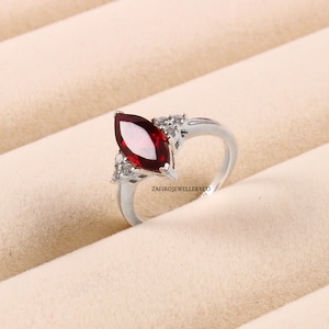 Puede incluir: Un anillo de plata con una gran piedra roja en forma de marquesa rodeada de piedras pequeñas y transparentes.