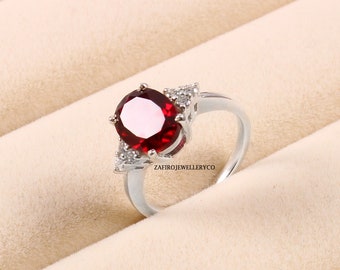 Puede incluir: Un anillo de plata con una gran gema roja oscura de corte ovalado como pieza central. El anillo está adornado con gemas más pequeñas y claras a cada lado de la piedra principal. La banda es lisa y pulida. El texto "ZAFIROJEWELLERYCO" es visible.