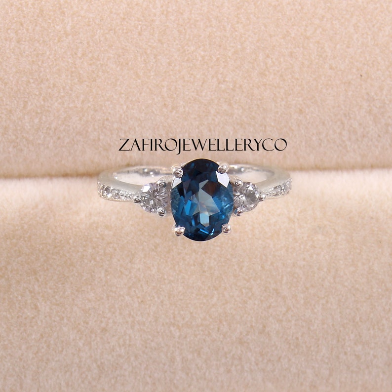 May include: A silver ring with a large oval blue gemstone and two smaller round white gemstones on either side. The ring is set in a white metal band.