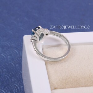 May include: A silver ring with a large blue oval gemstone surrounded by smaller clear gemstones. The ring is set in a white box with the text "ZAFIROJEWELLERYCO" printed on the side.