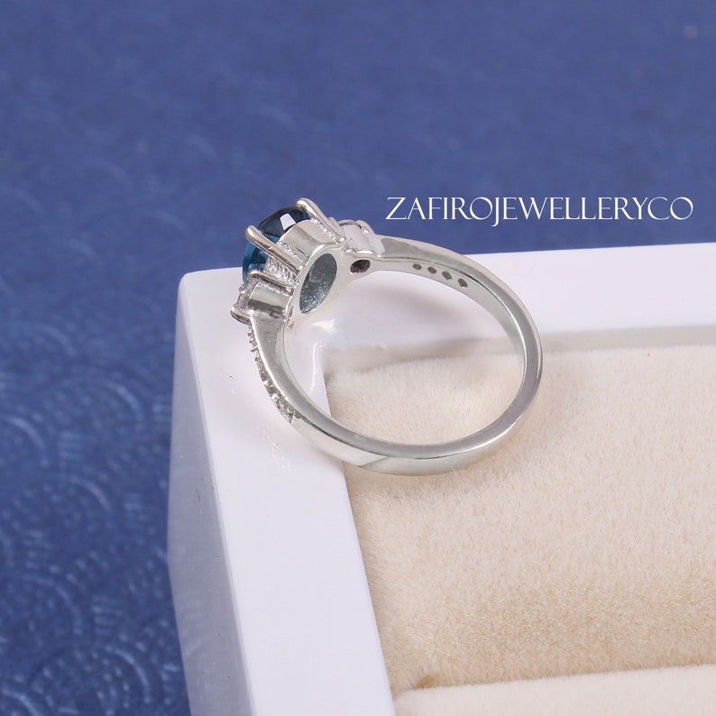 May include: A silver ring with a large blue oval gemstone surrounded by smaller clear gemstones. The ring is set in a white box with the text "ZAFIROJEWELLERYCO" printed on the side.