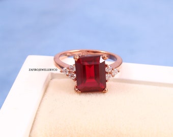 Könnte beinhalten: Roségoldener Ring mit einem großen, rechteckigen, tiefroten Edelstein. Kleine, klare Akzentsteine flankieren den Hauptstein. Der Ring ist vor einem hellblauen und weißen Hintergrund platziert. Der Text "ZafiroJewelleryCo" ist sichtbar.