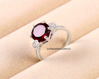 Peut inclure: Une bague en argent ornée d'une grosse pierre rouge foncé ronde. La bague est ornée de petites pierres précieuses claires de chaque côté de la pierre principale. Le nom de la marque "ZAFIROJEWELLERYCO" est visible sur la bague.