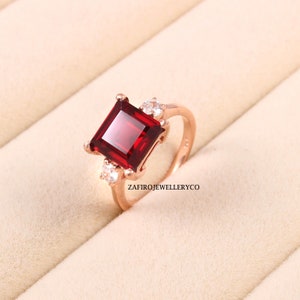 Puede incluir: Anillo de oro rosa con una gran gema roja oscura de corte cuadrado. Dos gemas claras más pequeñas flanquean la piedra principal. La banda del anillo es de oro rosa pulido. El texto "ZAFIROJEWELLERYCO" es visible.