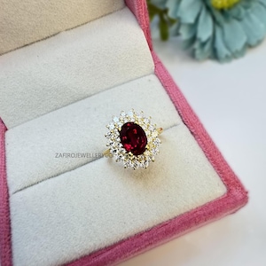 May include: A gold ring featuring a central oval-cut deep red gemstone surrounded by a halo of small clear stones. The ring is presented in a pink box with the text ZAFIROJEWELLERYCO visible on the white velvet insert near the jewelry piece.