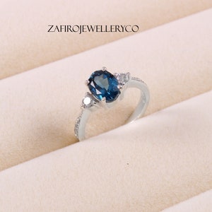 May include: A silver ring with a blue oval gemstone and two smaller white gemstones. The ring is set in a white metal band.