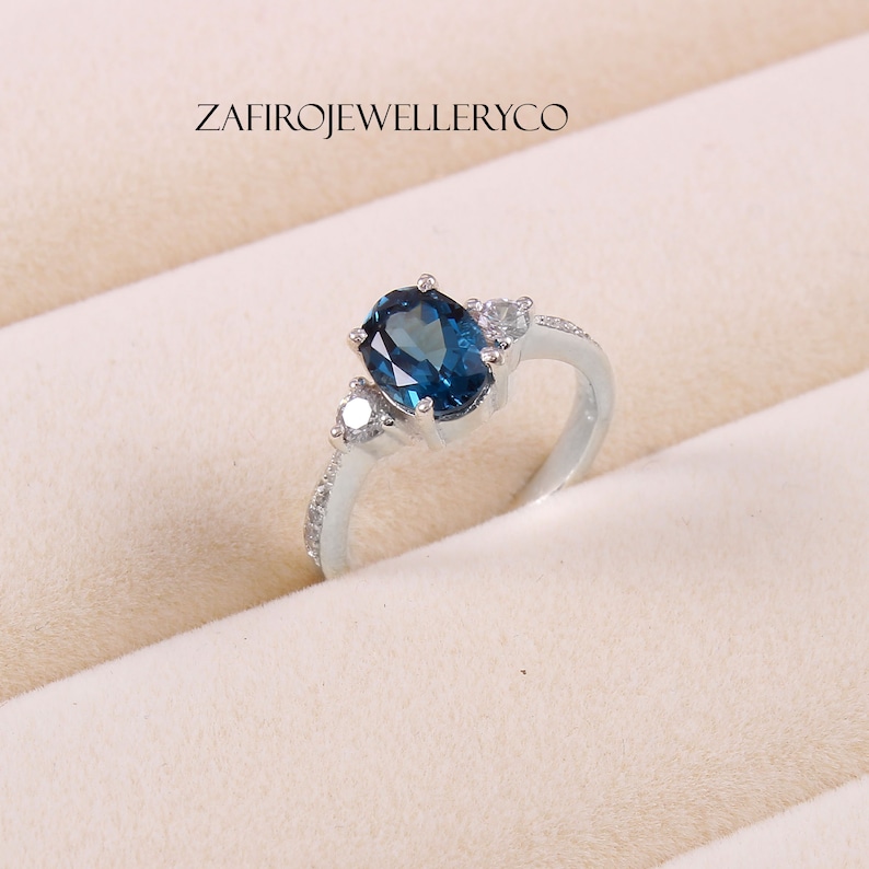 May include: A silver ring with a blue oval gemstone and two smaller white gemstones. The ring is set in a white metal band.