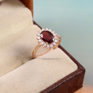 Peut inclure: Une bague en or rose avec une grosse pierre précieuse ovale rouge foncé, entourée de petites pierres claires. La bague est sertie dans un design en halo et est présentée dans un écrin marron. Le texte "ZAFIROJEWELLERYCO" est visible.