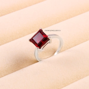 Peut inclure: Une bague en argent avec une grande pierre rouge de forme carrée. La bague est posée sur un fond blanc.