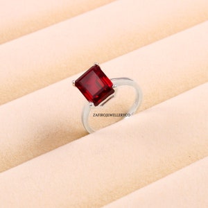 May include: A silver ring with a large, square, red gemstone. The ring is set on a white background.