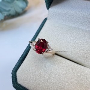 May include: A gold ring with a large, oval-cut, deep red gemstone as the focal point, complemented by smaller, triangular clusters of white gemstones. The ring is presented in a velvet-lined jewellery box.