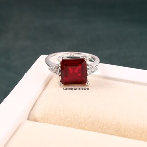 Peut inclure: Une bague en argent ornée d'une grande pierre rouge carrée. De petites pierres claires ornent les côtés de la pierre principale. La bague est présentée dans un écrin blanc. Le texte "ZafiroJewelleryCo" est visible.