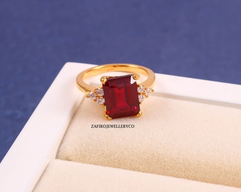 Könnte beinhalten: Ein goldener Ring mit einem großen, rechteckigen, tiefroten Edelstein als Mittelpunkt. Kleine, klare Edelsteine flankieren den Hauptstein. Der Ring ist vor einem blau-weißen Hintergrund platziert. Der Text "ZAFIRJEWELLERYCO" ist sichtbar.