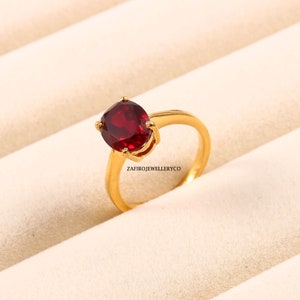 Peut inclure: Une bague en or avec une grosse pierre précieuse rouge foncé de forme ovale. La bague a un design simple et élégant avec un anneau lisse et poli. Le texte "ZAFIROJEWELLERYCO" est visible sur la bague.