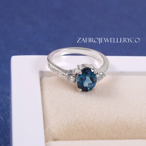 May include: A silver ring with a large blue oval gemstone and two smaller white gemstones on either side. The ring is in a white ring box.
