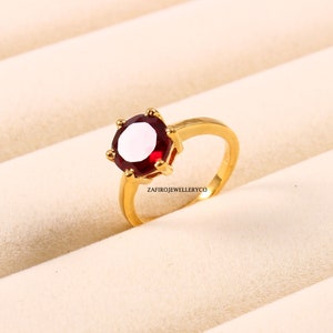 Könnte beinhalten: Ein goldener Ring mit einem großen, runden, tiefroten Edelstein. Der Ring hat einen schlichten Band und eine Fassung mit sechs Krappen. Der Markenname "ZAFIROJEWELLERYCO" ist auf dem Ring aufgedruckt.