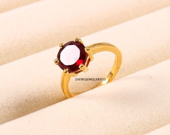 Peut inclure: Une bague en or avec une grosse pierre précieuse rouge foncé et ronde. La bague a un anneau simple et un sertissage à six griffes. Le nom de la marque "ZAFIROJEWELLERYCO" est imprimé sur la bague.