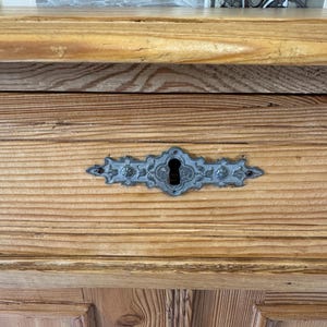 May include: Close-up of a wooden cabinet with a decorative metal keyhole plate. The cabinet is made of light-coloured wood with visible grain. The keyhole plate is a detailed, ornate design in a silver-grey colour.