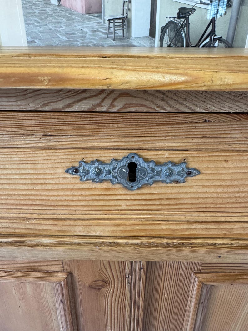 May include: Close-up of a wooden cabinet with a decorative metal keyhole plate. The cabinet is made of light-coloured wood with visible grain. The keyhole plate is a detailed, ornate design in a silver-grey colour.