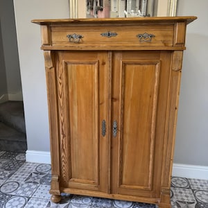 May include: A light brown wooden cabinet with two doors and a drawer. The cabinet features decorative metal handles and is positioned against a neutral wall. The cabinet is a piece of furniture.