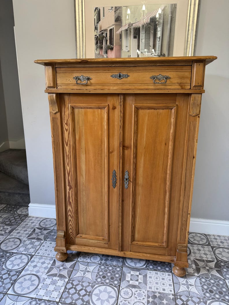 May include: A light brown wooden cabinet with two doors and a drawer. The cabinet features decorative metal handles and is positioned against a neutral wall. The cabinet is a piece of furniture.