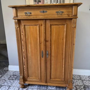 May include: A light brown wooden cabinet with two doors and a drawer. The cabinet has ornate metal hardware and is made of wood with visible grain. The cabinet is a vintage style piece.