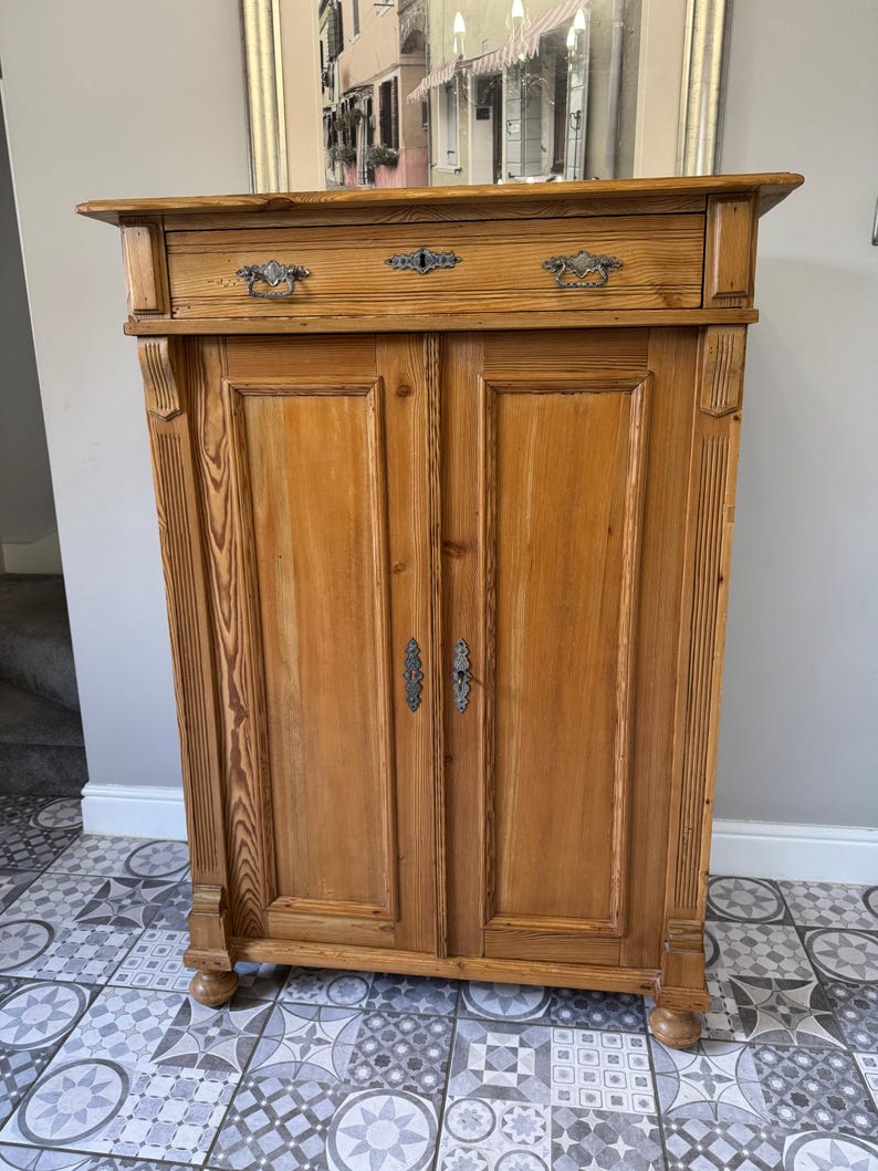 May include: A light brown wooden cabinet with two doors and a drawer. The cabinet has ornate metal hardware and is made of wood with visible grain. The cabinet is a vintage style piece.