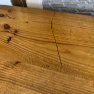 May include: Close-up of a section of a wooden surface with visible wood grain and knots. The wood has a warm, golden-brown colour. A thin, dark crack runs diagonally across the surface, adding a unique detail to the natural wood.