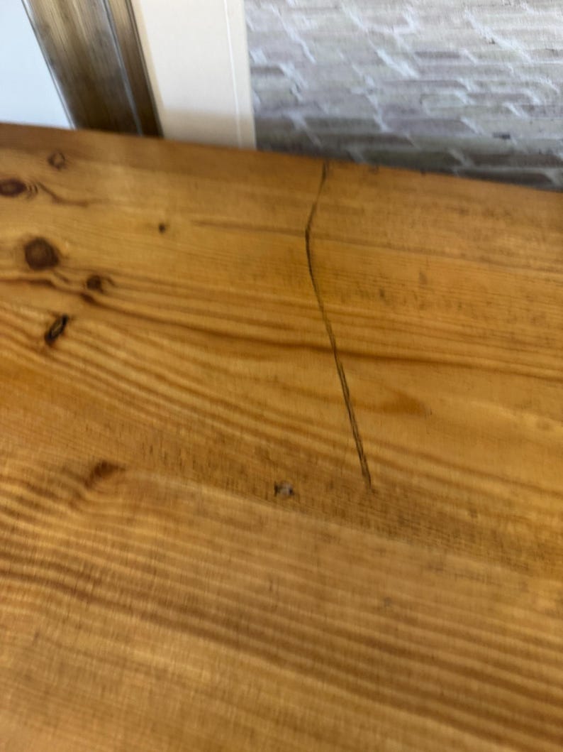 May include: Close-up of a section of a wooden surface with visible wood grain and knots. The wood has a warm, golden-brown colour. A thin, dark crack runs diagonally across the surface, adding a unique detail to the natural wood.
