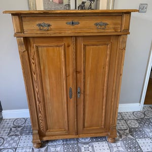 May include: A light brown wooden cabinet with two doors and a drawer. The cabinet has ornate metal handles and a keyhole. The wood grain is visible. The cabinet is on a patterned tile floor.